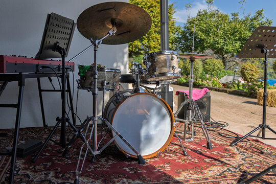 Acoustic drum kit and red stage keyboard stand on a patterned rug with music stands, cables and a sunny garden beyond the patio.