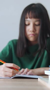 Vertical video close up and selective focus showing Asian kid girl concentrating as she writes in her notebook with a pencil. Bright daylight fills the room while she is doing her homework.