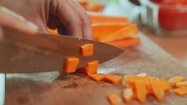 Woman hand dicing carrot with kitchen knife on wooden cutting board.