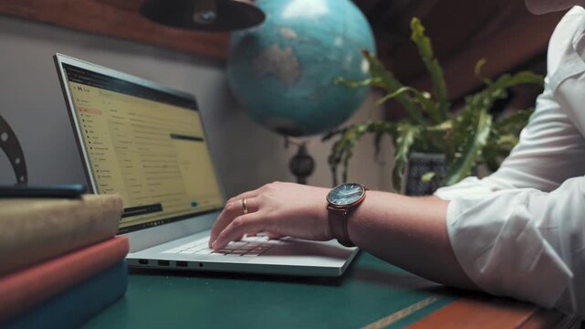 Businessman types on a slim silver laptop keyboard while sitting at his cluttered desk with stacked books and a globus nearby in an old study room that has soft lighting and some empty copy space.