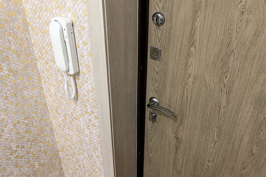 beige wood‑grain entry door with a metallic lever handle, cylinder lock, and plate. left, a white wall‑mounted intercom handset rests on patterned wallpaper