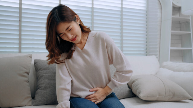 An Asian young woman sitting on sofa and holding her painful stomach, suffering from severe abdominal pain, menstrual cramps, food poisoning, indigestion, or gastritis symptom at home living room