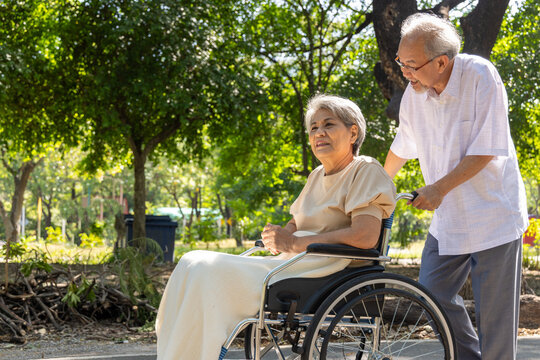 An Asian senior man lovingly pushes his elderly wife in a wheelchair through a sunny park, providing warm assistance, compassionate caregiving, and dedicated support