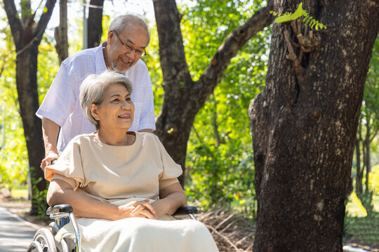 An Asian senior man lovingly pushes his elderly wife in a wheelchair through a sunny park, providing warm assistance, compassionate caregiving, and dedicated support