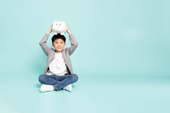 A cheerful Asian boy sitting cross-legged with a white piggy bank on his head in studio, illustrating money saving, childhood financial planning, and education fund concept with copy space