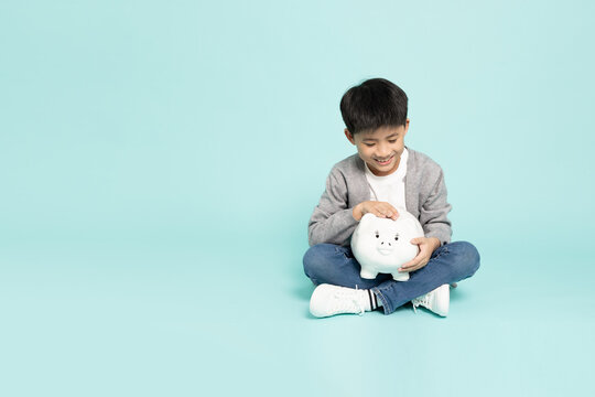 A happy Asian boy sitting cross-legged putting coin into white piggy bank in studio, showcasing money saving habit, child financial literacy, and education investment with copy space