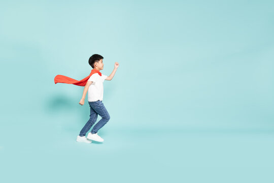 A brave Asian boy in a red superhero cape running forward with determination in studio, representing child imagination, power, courage, and leadership with empty copy space on blue background