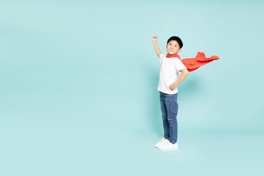 A brave Asian boy wearing a red superhero cape raising his fist with confidence in studio, portraying child strength, power, leadership, and success concept on light blue background with copy space