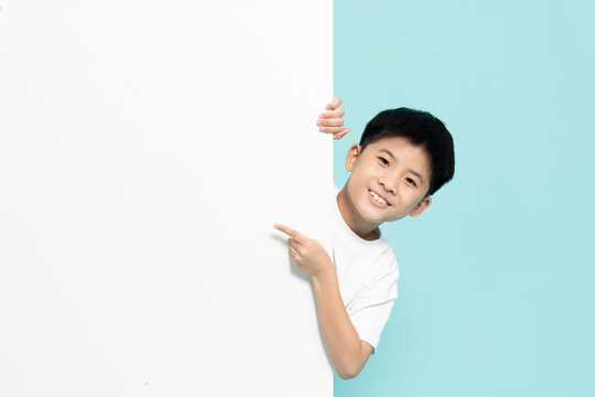 A happy Asian boy peeking behind a blank white banner board and pointing finger at empty copy space for advertisement, marketing presentation, and promo concept on light blue background