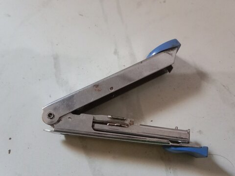 This image shows a silver metal stapler with blue plastic handles in an open position on a white surface. The stapler appears to be used, with minor rust spots and scratches visible on the metal body.