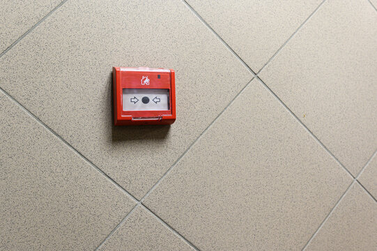 red manual fire alarm button mounted on a beige tiled wall, featuring directional arrows, a central actuator, and a house-shaped safety icon above the activation panel