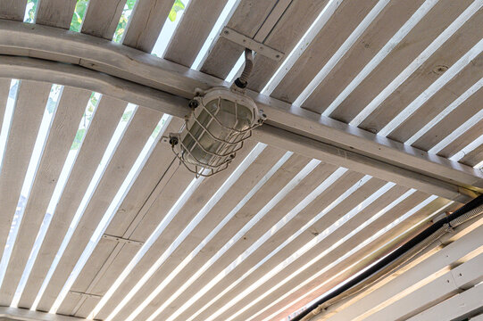vintage-style industrial light fixture with a wire cage mounted on a curved white beam under a slatted wooden ceiling. visible conduits and fasteners complete rustic outdoor look