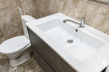 Fototapeta premium compact bathroom with a white rectangular sink atop a grey wood-grain vanity. chrome faucet, white toilet, and beige marble-patterned tiles complete the setup