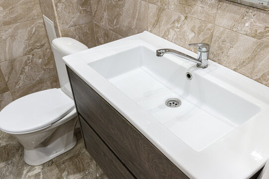 compact bathroom with a white rectangular sink atop a grey wood-grain vanity. chrome faucet, white toilet, and beige marble-patterned tiles complete the setup