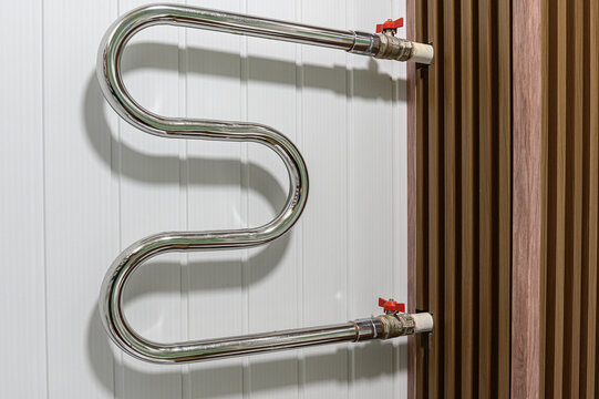 chrome‑plated S‑shaped heated towel rail mounted on a white paneled wall. connects to a radiator via pipes with red valve handles
