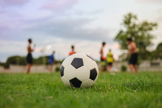 Soccer ball on green grass field with blurred silhouette of children playing football at sunset, golden hour light. Kids sport training concept