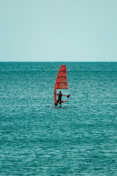 A windsurfer glides across the calm sea with an orange sail under an overcast sky.