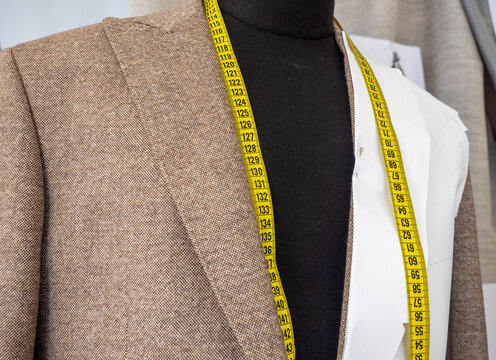 A brown tweed jacket in progress with paper pattern template and yellow measuring tape draped on a black tailor mannequin in an atelier.