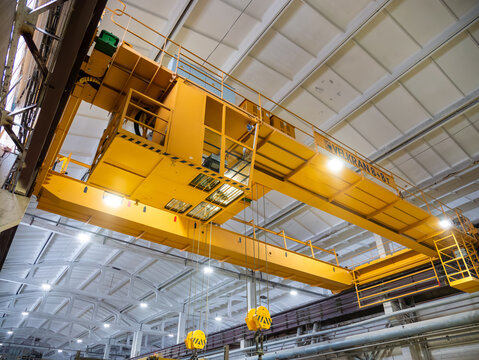 A large yellow overhead gantry crane with heavy lifting hooks operates beneath the ceiling structure of a modern industrial factory plant.