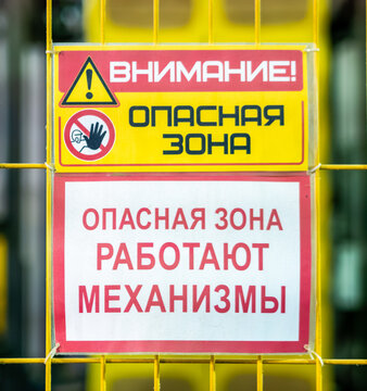 Russian language industrial safety warning signs attached to a yellow protective metal grid fence warn of a danger zone and operating machinery.