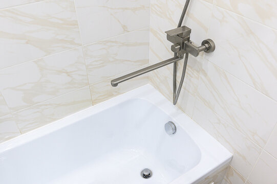 clean white bathtub in a corner with beige marble‑effect tiles. brushed‑metal mixer tap, shower arm, and flexible hose are mounted on the wall