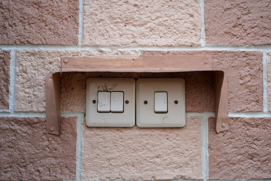 Vintage light switches mounted on textured pink brick wall with metal bracket, showing aged electrical fixtures in a rustic home setting, featuring intricate old details.