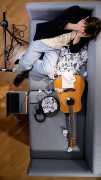 Frustrated musician covering face with hands, lying on a sofa surrounded by crumpled paper, a guitar, laptop, and microphone, struggling with writer's block in a home studio
