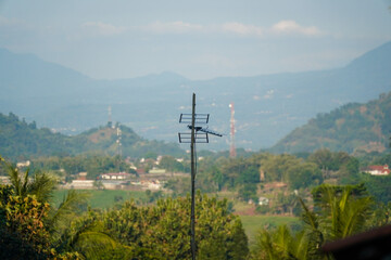 Fototapeta premium A weathered television antenna stands tall against a lush green valley with distant mountains and scattered villages under a hazy sky, capturing rural connectivity amidst nature.