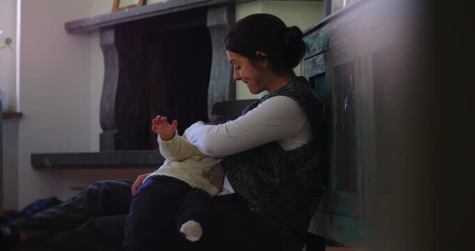 Mother breastfeeds young child while seated on floor and leaning against wooden cabinet at home, expressing maternal nourishment, comfort, attachment and intimate bonding