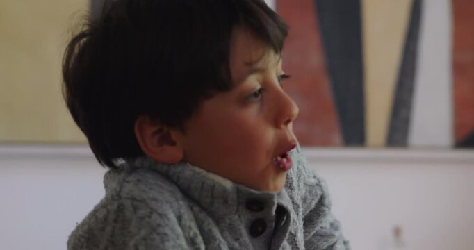 School-age boy eats coconut pastry while looking away in home dining room, savoring a sweet snack with quiet enjoyment, independence and everyday childhood contentment