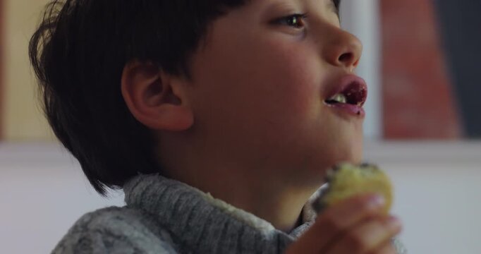 Young boy bites coconut-covered cake while watching someone nearby at home dining table, enjoying a casual dessert break, sweet reward and relaxed childhood moment