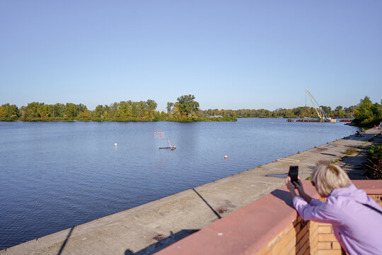 A woman leans on a brick railing to photograph a calm river and tree-lined shoreline with a distant crane, capturing a peaceful waterfront scene on a clear sunny day.
