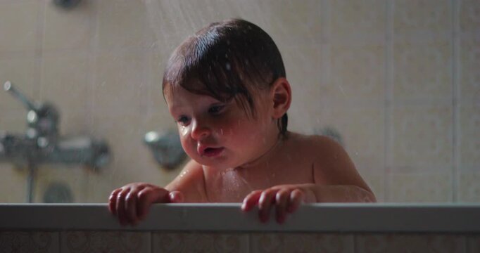 Curious toddler peers over bathtub rim while shower water falls in home bathroom, experiencing cautious sensory discovery, playful exploration and childhood learning