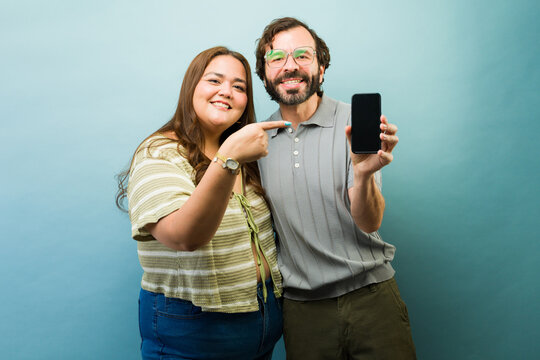 Smiling couple presenting smartphone with blank screen, woman pointing, recommending new app or online service
