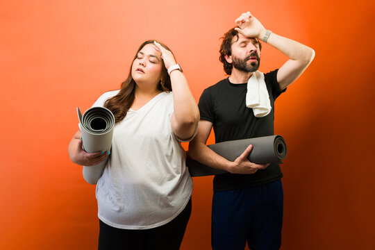 Exhausted plus size woman and man wiping sweat after a fitness workout, holding yoga mats with a vibrant orange background