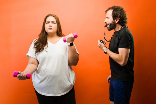 Overweight woman looking intense during personal training session, male coach shouting instructions and timing exercise