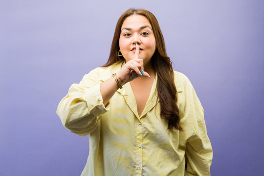 Curvy woman hushing with finger on lips, asking for silence and secrecy on purple background