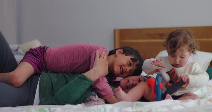 Mother cuddles older son on bedroom bed while toddler plays nearby with toy guns, capturing sibling contrast, divided attention and authentic everyday parenting
