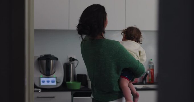 Tired mother yawns while holding toddler in home kitchen, illustrating maternal fatigue, childcare multitasking, mental load, overwhelm and everyday parenting demands