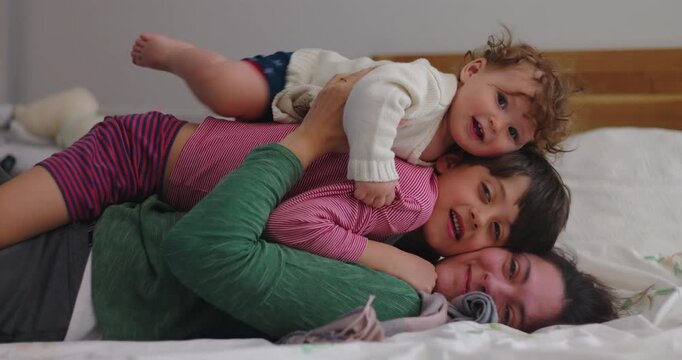 Mother lies under two sons piled on her during bedroom roughhousing, balancing playful chaos, maternal patience, physical affection and authentic family bonding
