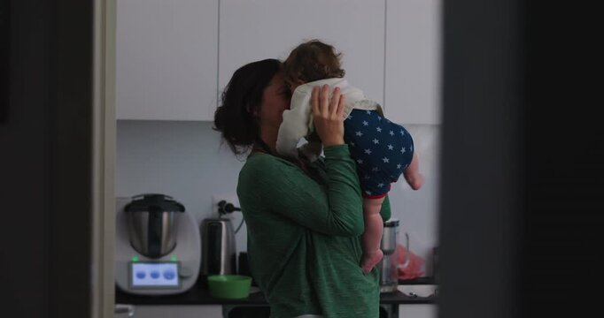 Mother holds toddler as child leans in to kiss her in home kitchen, sharing reciprocal affection, playful attachment, family love and authentic joyful bonding