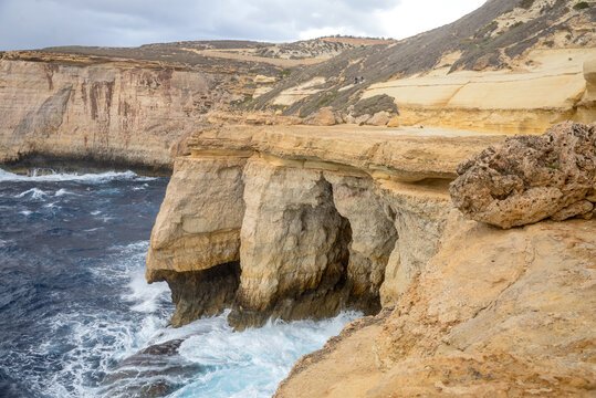Rugged limestone cliffs and sea caves on the Gozo coastline in Malta, featuring crashing blue waves under a cloudy sky.