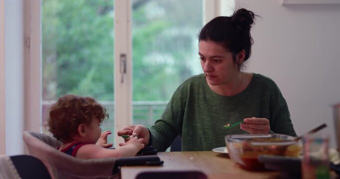 Mother spoon-feeds toddler at home dining table, smiling and encouraging child during family meal, expressing responsive caregiving, nutrition and joyful connection