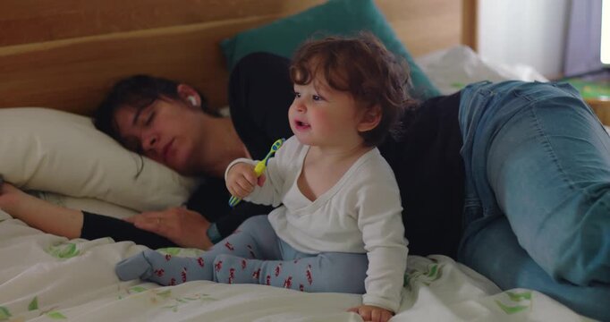 Toddler sits on bedroom bed holding toothbrush and pretending to brush through imitation while mother sleeps deeply, revealing learning, maternal fatigue and overwhelm