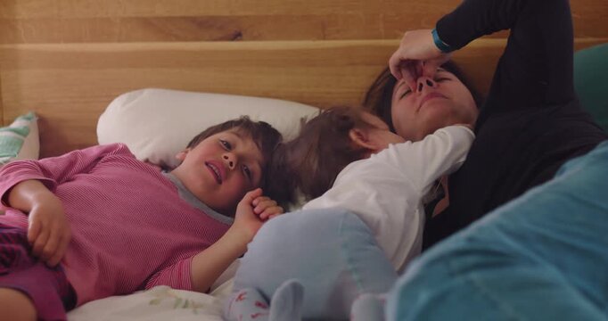 Siblings play energetically on bedroom bed while overwhelmed mother tries to rest behind them, illustrating maternal fatigue, mental load and authentic family chaos