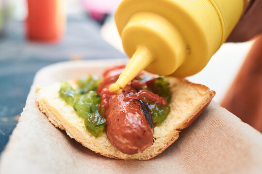 Mustard being squeezed from a yellow bottle onto a hot dog with relish and ketchup