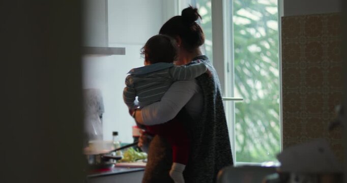 Mother carries toddler while cooking at stove in home kitchen, balancing meal preparation, childcare, household labor, multitasking demands and devoted everyday parenting