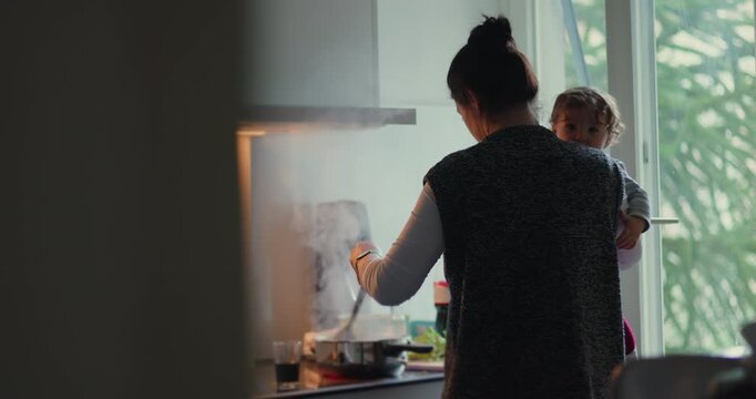 Busy parent stirs pot while holding young child on her hip in home kitchen, combining family cooking, childcare, mental load and modern caregiving responsibility