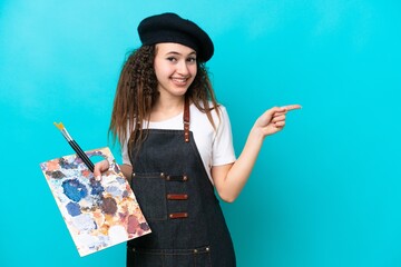 Young artist Arab woman holding a palette isolated on blue background pointing finger to the side © luismolinero