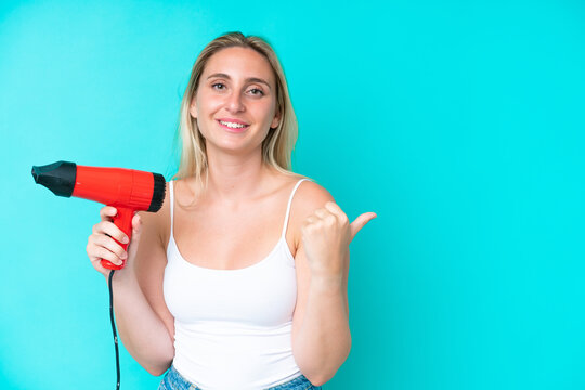 Young caucasian woman holding a hairdryer isolated on blue background pointing to the side to present a product